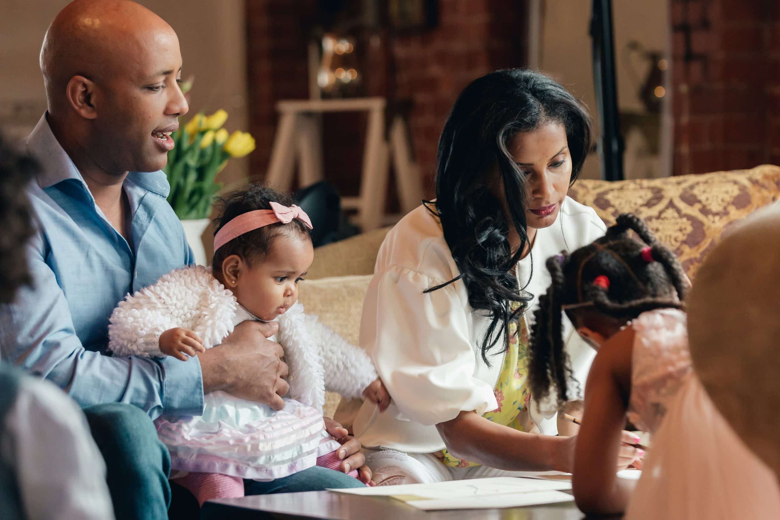 parents-and-two-daughters-in-family-room A family at home: a father holding a baby while the mother helps a young girl draw at the table